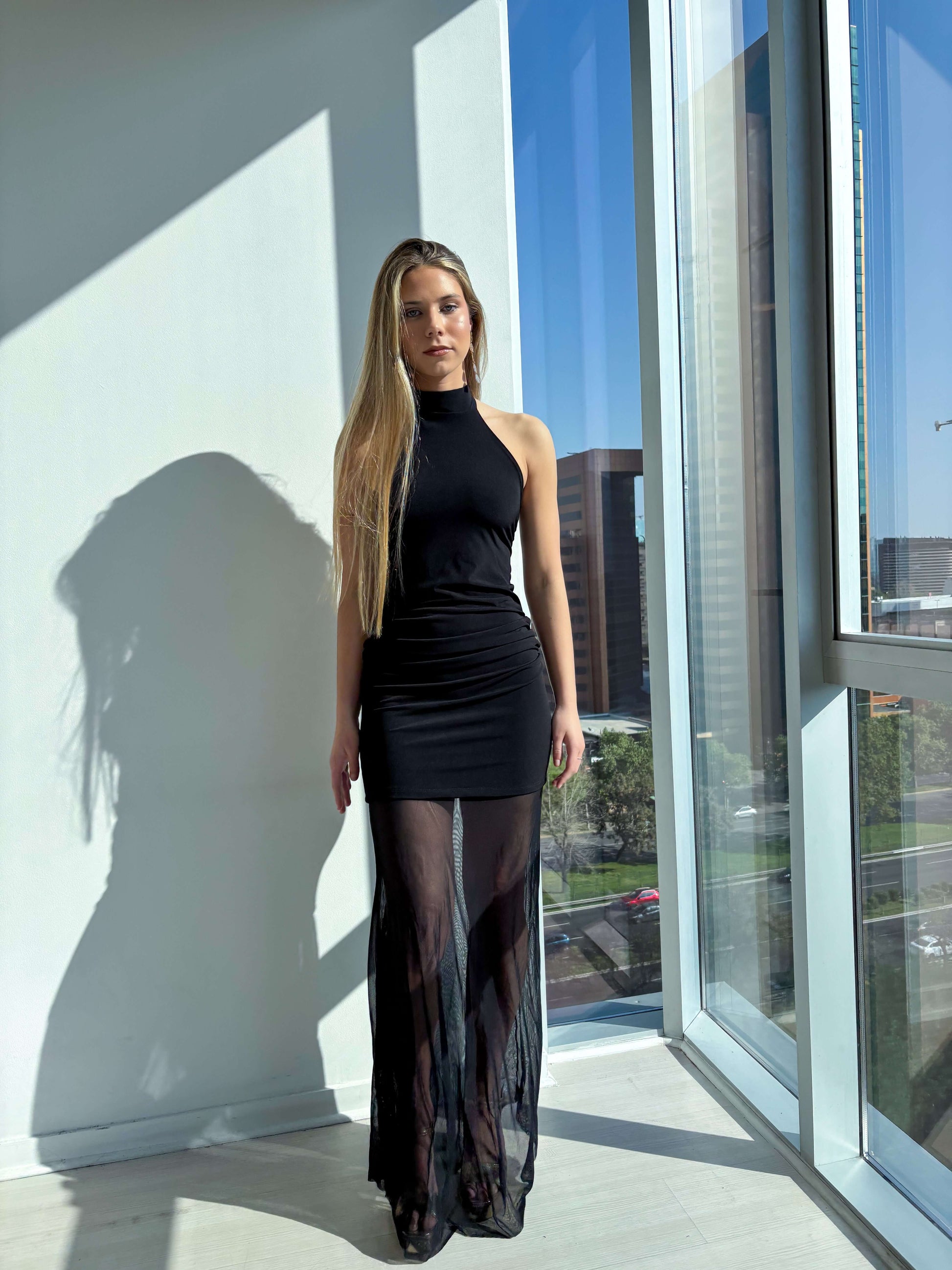 Modelo luciendo vestido elegante negro con cuello halter y falda de transparencia en ambiente moderno.