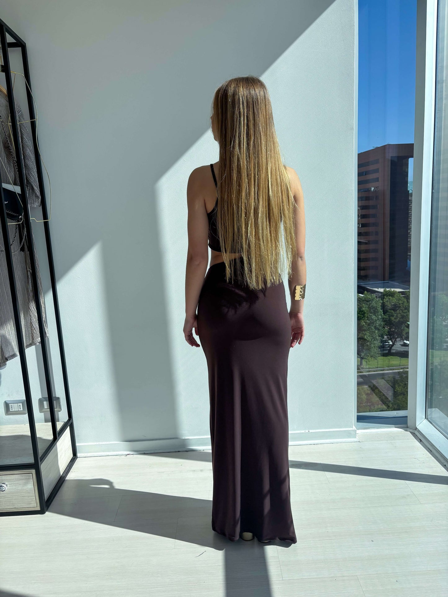 Mujer de espaldas luciendo el vestido de fistes asimetrico cafe hacia la ventana iluminada.